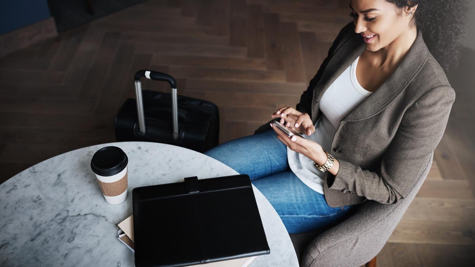 Ein Smartphone mit einem geöffneten Chatfenster liegt auf einem Holztisch neben einer Kaffeetasse in einer Hotellobby