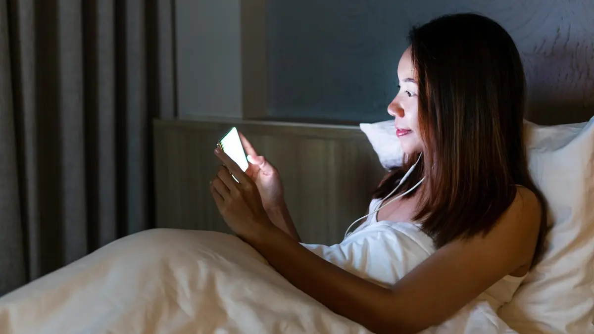 Smartfon z otwartym czatem leżący na drewnianym stoliku nocnym w pokoju hotelowym obok włączonej lampki