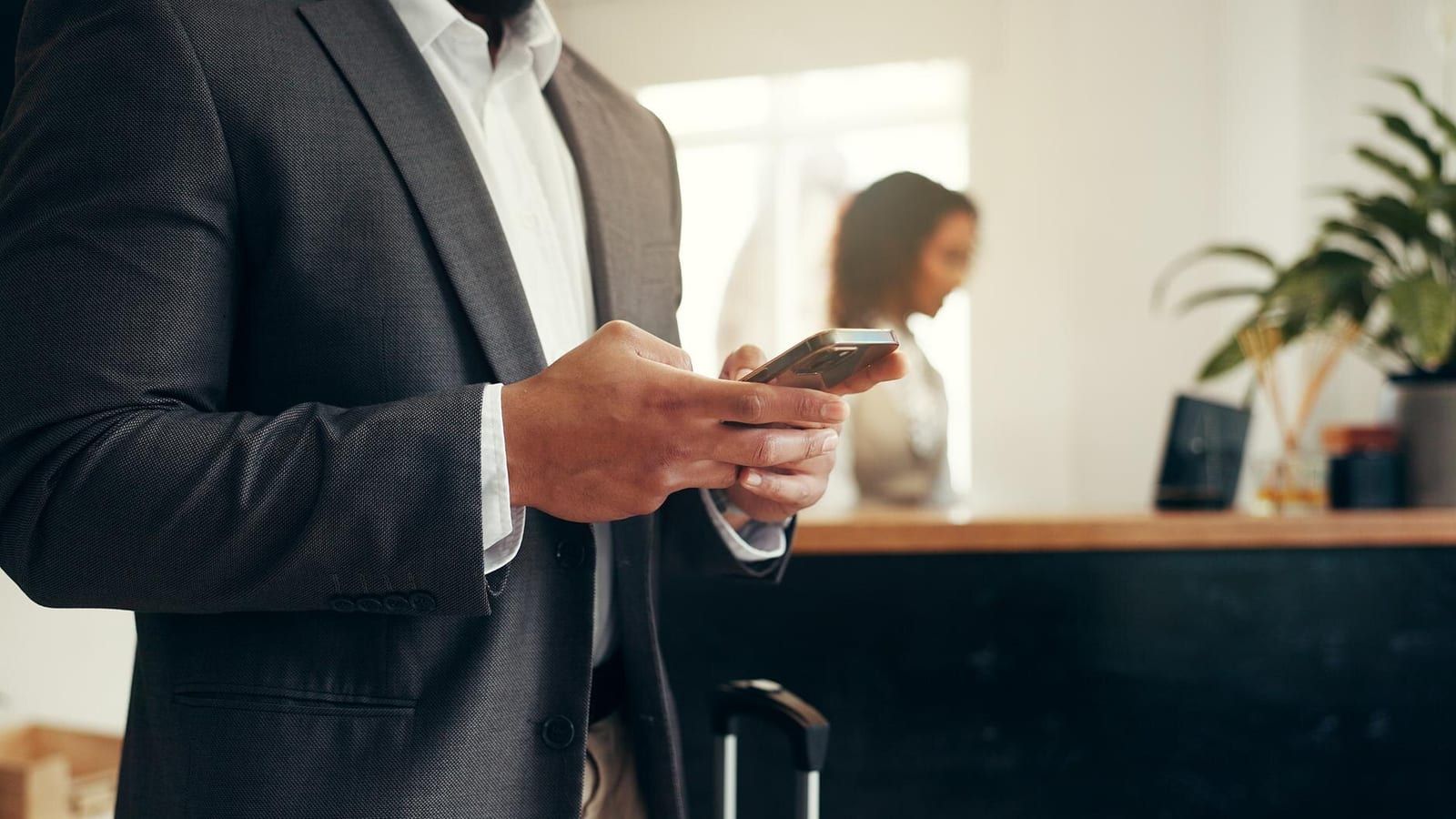 Ein Hotelgast tippt auf seinem Smartphone an einem Holztisch in der Lobby, im Hintergrund eine unscharfe Rezeption