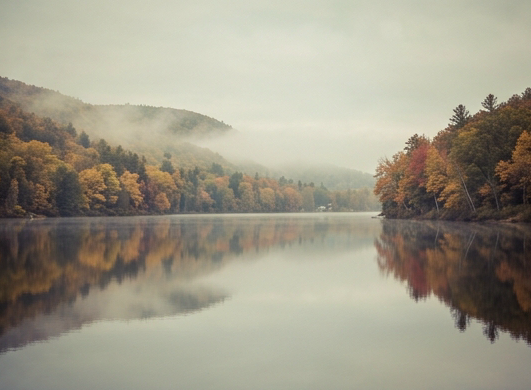 Landscape photograph of Otsego Lake near Cooperstown New York, calm glassy glacial water reflecting autumn foliage, forested hillsides in gold and red, misty morning atmosphere, muted desaturated vintage photographic tone, wide angle, serene and pristine natural beauty