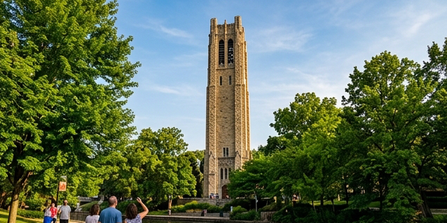 Photograph of Moser Tower and Millennium Carillon in Naperville Illinois, a tall 160-foot stone tower rising dramatically above green trees along the riverwalk, warm summer afternoon light, blue sky backdrop, visitors looking up at the tower, editorial travel photography with vibrant greens and natural stone textures.